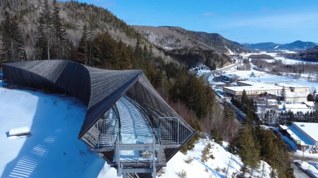 Aerial view of the curved wooden observation walkway at Belvedere des Deux-Rivieres. Panoramic drone shot overlooking a snowy valley, coniferous forest, and the town of Matapedia in Quebec.