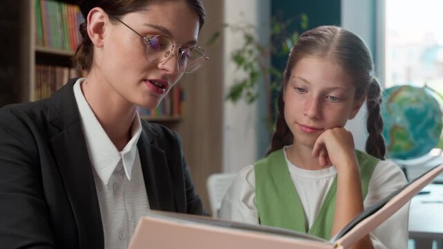 Caucasian personal tutor female teacher woman reading story book literature course read textbook to little girl child kid schoolkid pupil listening lesson listen in school elementary primary education