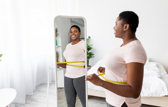 Overweight young black woman measuring her waist with tape near mirror at home, satisfied with slimming result, free space. Plus size African American lady being successful in weight loss