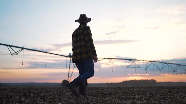 Farmer walking in field with irrigation system at sunset. Rural farmer inspecting crops and water supply. Agricultural lifestyle focused on field irrigation. Farmer in boots hat managing water system.
