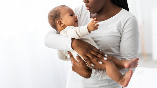 Portrait of young African American mother holding her crying baby on hands. Displeased hungry infant whining, feeling pain or colic. Copy space, blurred background. Motherhood And Maternity Concept