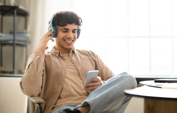 Glad Young Arab Guy Having Fun With Smartphone, Listens Online Music In Wireless Earphones, Sitting Near Desk During Work Day At Home Interior. Modern Gadgets Lifestyle Concept
