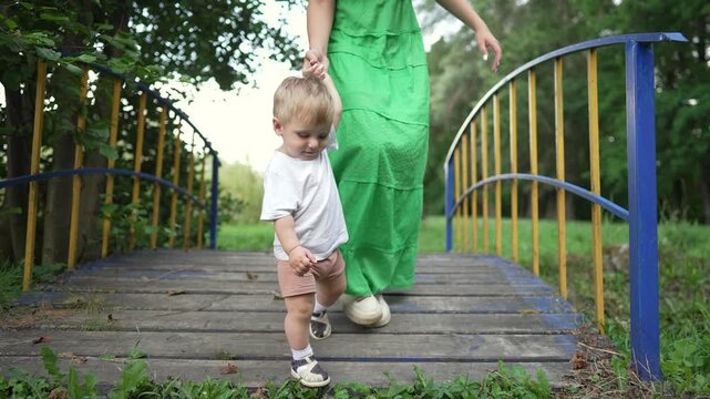 Mother helps toddler take first steps. Family bonding in nature. Toddler learns to walk outdoors. First steps on grass with family. Mother and child in nature. Toddler first learning experience.