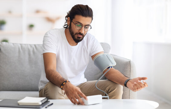 Young arab man sitting on couch, checking blood pressure and taking notes at home. Indian guy in white t-shirt using modern tonometer, experiencing hypertension caused by stress, copy space