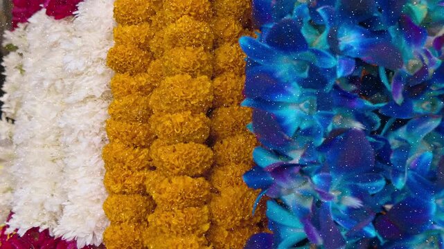 Beautiful natural fragrant flowers - orange marigolds, blue Dendrobium orchids and white flowers gathered into beautiful garlands for offerings and wedding purposes in India. View through sparkling pa