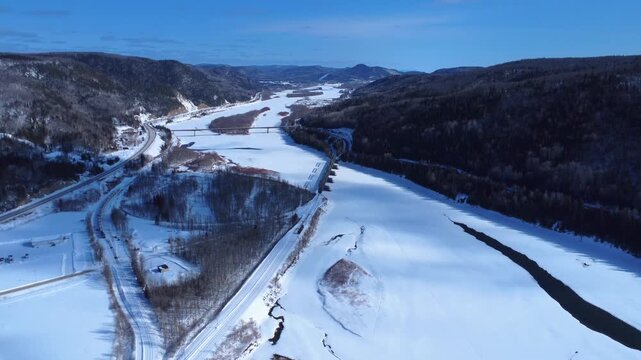 Panoramic drone shot of the Restigouche Salmon River in a snowy valley. Features railway and highway bridges at the natural border between Quebec and New Brunswick in a winter mountain landscape.