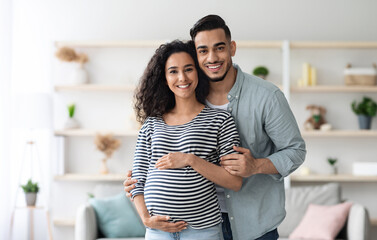 Fototapeta premium Portrait of happy arabic expecting family posing together at home, handsome middle eastern millennial man and beautiful long-haired pregnant lady embracing and smiling at camera, copy space
