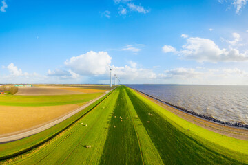 Obraz premium Aerial view of wind turbines on green fields near sea coast and dam. Scenic landscape with wind generators, meadow, ocean and cloudy sky at sunset. Renewable energy and environment from above.