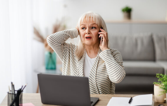 Oh No. Portrait of shocked frustrated elderly woman having problems with computer or work, talking on cellphone with customer support while working in home office grabbing head