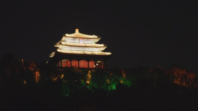 Distant illuminated pavilion above treeline, small lit pavilion rises above dark canopy, golden roof silhouette contrasted against black sky, surrounding trees washed with subtle colored uplighting,