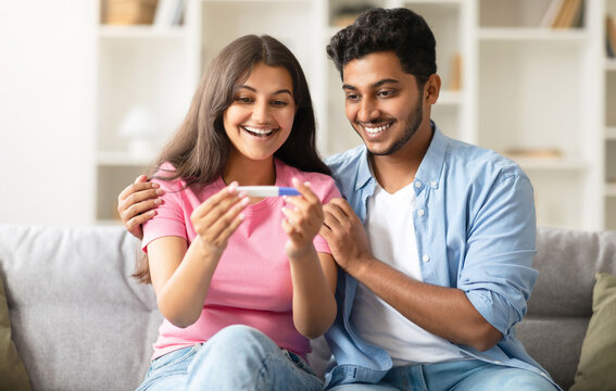 Excited indian couple looking at positive pregnancy test, smiling and enjoying great news, sitting on couch at home. Loving hindu spouses feeling happy about future parenthood