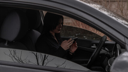 Naklejka premium woman driving a black car with a phone in her hands. black dark hair, makeup, open window, driving, studying, texting, working, talking