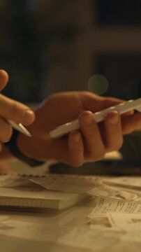 A man leans over his calculator, eyes fixed as he flips through receipts and scattered papers on a cluttered desk. The dim, softly lit room hints at a late-night quiet budgeting session