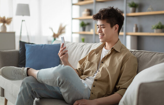 Cheerful Young Asian Man Using Smartphone Texting Online And Scrolling News Feed In Social Media Application Sitting On Couch At Home. Happy Guy Reading Message On Cell Phone. New Mobile App Concept