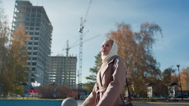 Woman with headscarf walking construction site bathed in crisp autumn light, wearing beige coat and shoulder bag, passing crane and unfinished highrise beside golden trees under clear blue sky, candid