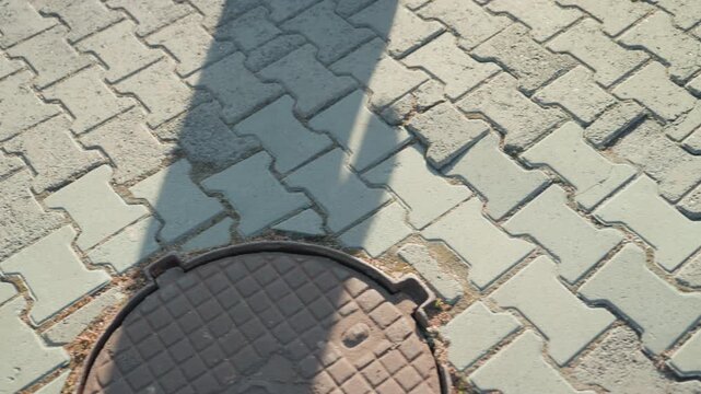 Long shadow across patterned pavement pink coat and boots, silhouette stretching over manhole cover, contemplative commuter slowing step, cinematic angle emphasizing geometry and tile texture, quiet
