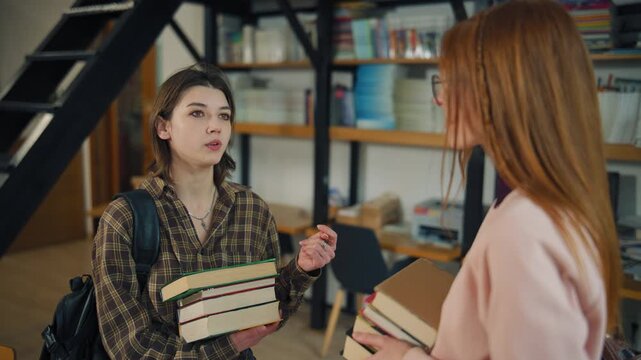 Two students discuss and share books in a library during the day while standing near bookshelves filled with more books