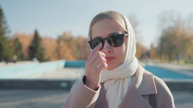 White woman adjusting sunglasses urban plaza, assertive expression with pointing gesture, layered coat and scarf, golden sunlight and crisp leaves, influencer vibe with cinematic backlight, candid