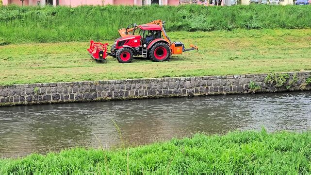 A professional red tractor equipped with a hydraulic arm flail mower performs seasonal grass cutting on a steep river canal slope.