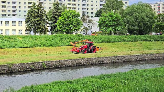 A professional red tractor equipped with a hydraulic arm flail mower performs seasonal grass cutting on a steep river canal slope.