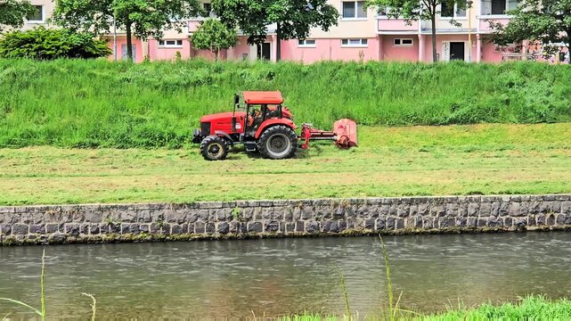 A professional red tractor equipped with a hydraulic arm flail mower performs seasonal grass cutting on a steep river canal slope.