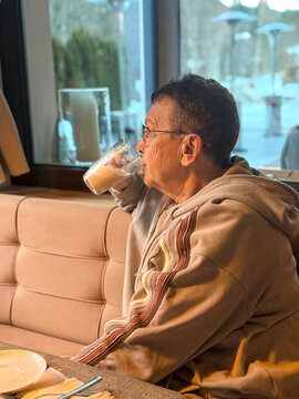 A senior woman sits at a table, drinking coffee while looking out the window. Snowy weather is visible outside. She wears a warm outfit and seems to enjoy her drink.