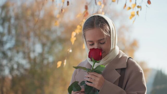 Young woman with scarf smelling rose in autumn park, romantic recipient pausing under birch branches, contemplative portrait with soft sunlight and golden foliage, cozy coat, delicate petals, serene
