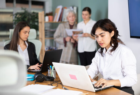 Asian girl sitting at desk is working on laptop computer among staff colleagues. Working day in office of business structure, manager is working at computer.
