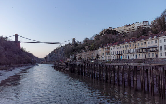 A view of the Clifton Suspension Bridge in Bristol, England, spanning the Avon Gorge, with rows of houses on the hillside and a river in the foreground.
