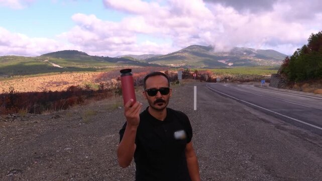 Slow motion of a man standing by a highway next to a broken car and showing a thermos while making an okay gesture with his hand for roadside rest concept
