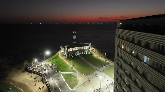  Aerial reveal of Farol da Barra lighthouse emerging from behind a coastal building, with illuminated shoreline, beach, and ocean under a red dusk sky.
