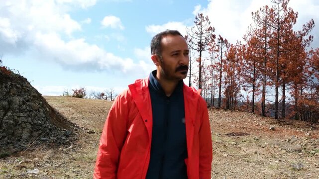Man in a vibrant orange waterproof jacket walking alone through a devastated forest landscape with burnt trees and black ash soil after a natural disaster at daytime