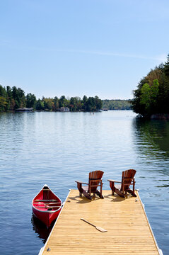 Two Adirondack chairs and a red canoe on a sunny wooden dock in cottage country Ontario, Canada. Paddleboarders enjoy a calm summer lake, vacations, retirement, beauty in nature, tranquility.