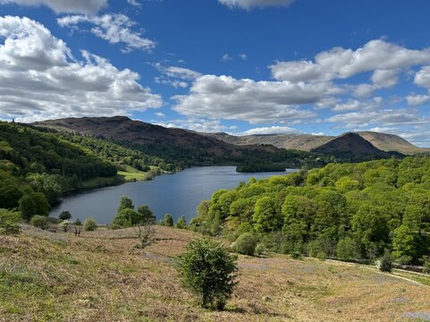 Panoramic view of Grasmere Lake with calm water, lush trees and surrounding hills beneath a partly cloudy blue sky. Lake District, England, UK.
