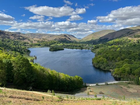 Wide view of Grasmere Lake with calm water, green trees and surrounding hills under a partly cloudy blue sky. Lake District, England, UK.