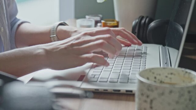 Hands typing on laptop keyboard at desk with mug, headphones and plant. Concept of remote work, home workspace and productivity