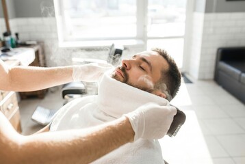 Fototapeta premium Barber applying a steaming hot towel to the face of a male client in a barbershop