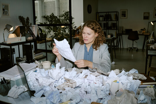 Caucasian young woman sitting at cluttered office desk reading crumpled paper surrounded by piles of discarded documents and paperwork in modern business workspace