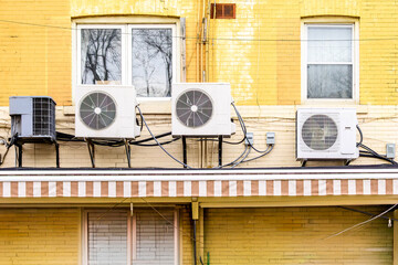 Fototapeta premium Three mini-split air conditioner condenser units installed side by side on exterior ledge of yellow brick multi-unit building.