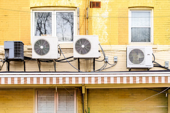 Three mini-split air conditioner condenser units installed side by side on exterior ledge of yellow brick multi-unit building.