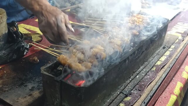 Closeup of a hand grilling chicken satay skewers over hot coals with smoke rising a popular street food in Southeast Asia. 4k Footage Video