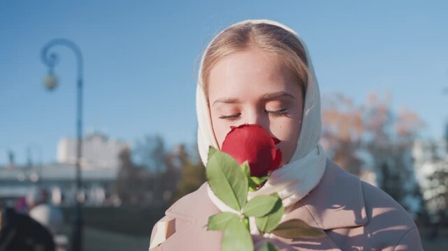 White woman smelling red rose city, young white woman in scarf pauses on riverside promenade under winter sunlight, closes eyes to inhale rose, soft expression, lamppost and skyline in background,