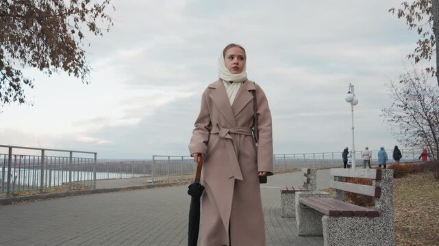 White woman posing with umbrella, beige trench coat and boots, editorial model stance and calm gaze, waterfront backdrop, autumn trees and cloudy sky, closeup portrait framing, elegant leather bag,