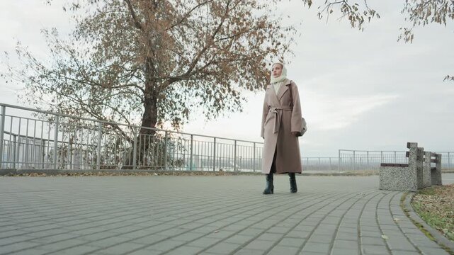 Woman coat scarf walking promenade city, park trees with fallen leaves and bench near metal railing, waterfront panorama and distant skyline, crisp chilly air and purposeful stride