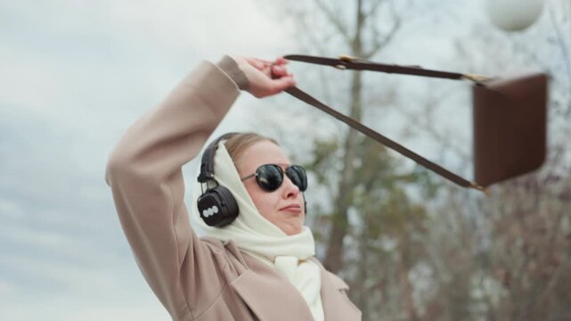 White woman park playful music dance tossing purse and laughing with headphones and sunglasses, leafless trees and cloudy sky, candid energetic motion and modern street fashion vibe