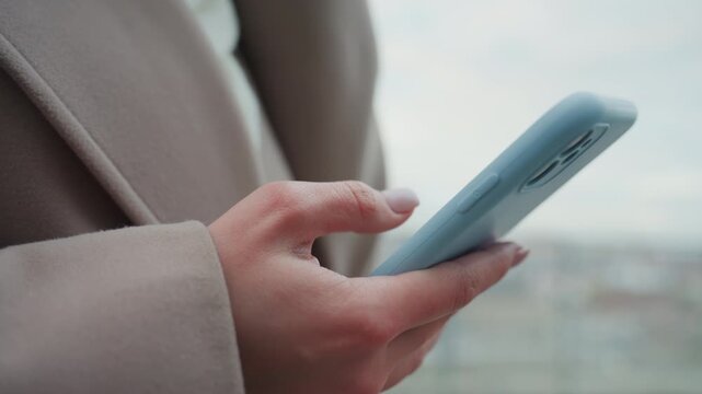 Hands holding blue phone outdoors, adult commuter in beige coat taps screen on cold overcast morning with blurred city skyline in background, closeup detail of fingers scrolling and interacting