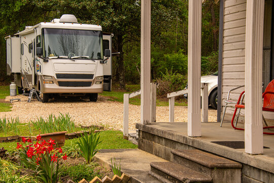 A camper van is parked in a rustic setting beside an old wooden house. The scene is framed by vibrant garden flowers and a stone porch.