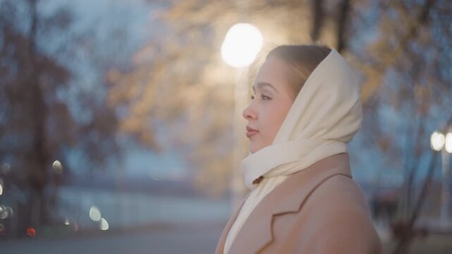 Woman with shawl walks through park darkness. Young lady stands calmly under glowing street lamps in park. Young woman wearing scarf pauses peacefully beneath streetlights in park during dusk
