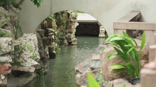 Tranquil pond under stone arch, lush green plant in foreground, moss covered rocks bordering calm water, gentle ripples reflecting archway, secluded courtyard atmosphere with soft sunlight and shadow,