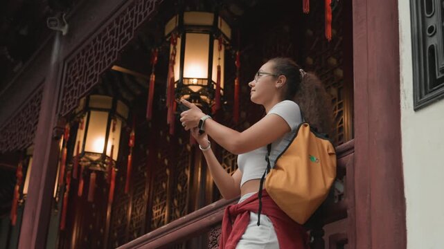 Asian woman photographing temple lanterns with smartphone wearing yellow backpack, framing glowing lantern cluster under ornate eaves travel photographer vibe, eager curiosity, market ambiance,